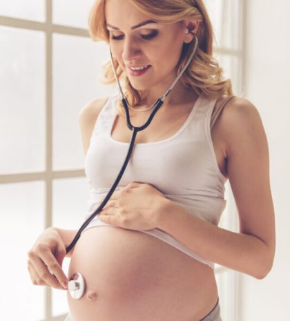 Beautiful pregnant woman is listening to her tummy using a stethoscope and smiling